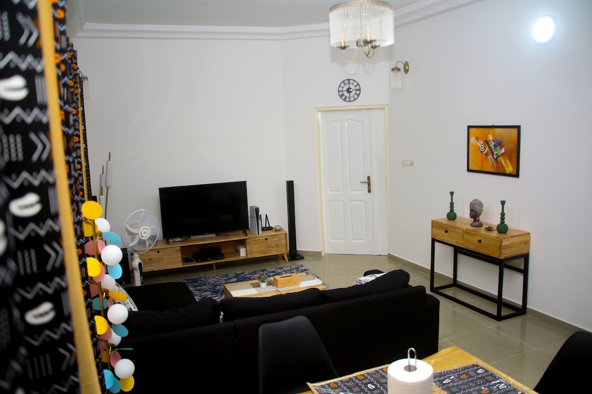 A living area features a modern black sofa and a wooden media console with a flat-screen television. Decorative elements include a round wall clock and two vases atop a side table. A dining area is partially visible, adorned with a patterned tablecloth and a light fixture overhead.