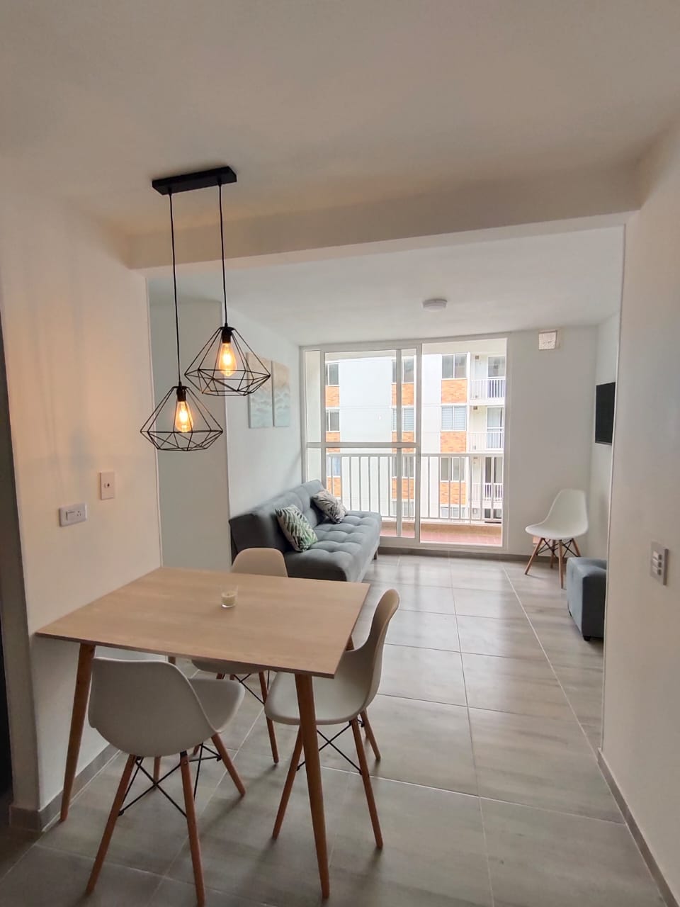 A well-lit dining area features a small table surrounded by four white chairs. Two industrial-style pendant lights hang overhead, adding a modern touch. A comfortable gray sofa is positioned nearby, with large windows allowing natural light to fill the space.