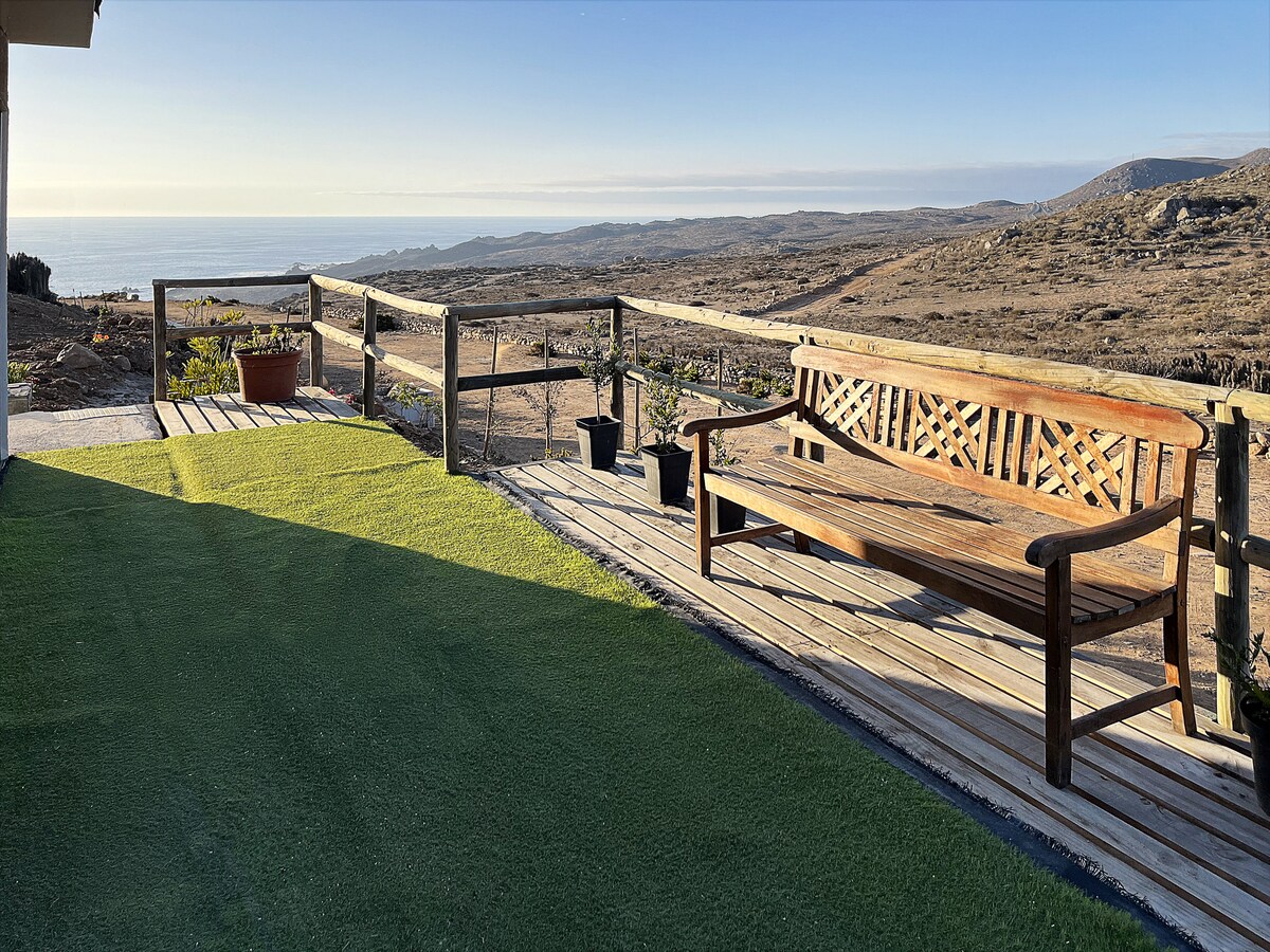 A wooden bench is positioned on a spacious deck overlooking a serene coastal view. The landscape features arid terrain stretching to the ocean, with potted plants enhancing the area. The early morning light casts soft shadows across the green carpeted surface.