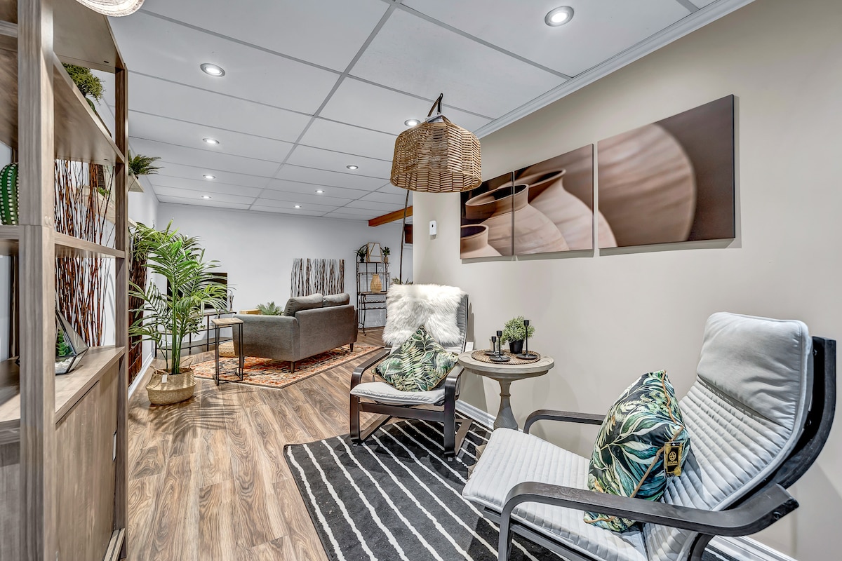 A cozy lounge area in the basement features two comfortable armchairs adorned with decorative pillows. A modern shelf displays various plants and decorative items. Soft overhead lighting enhances the inviting atmosphere, while a patterned rug provides warmth to the space.