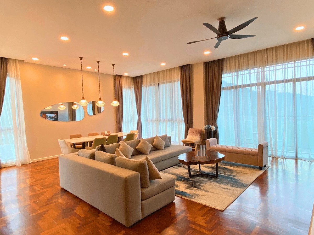 A spacious living area is highlighted by large windows allowing natural light to fill the room. Comfortable seating is arranged around a coffee table, with a dining area visible in the background. Soft curtains drape beside contemporary light fixtures, enhancing the inviting atmosphere.