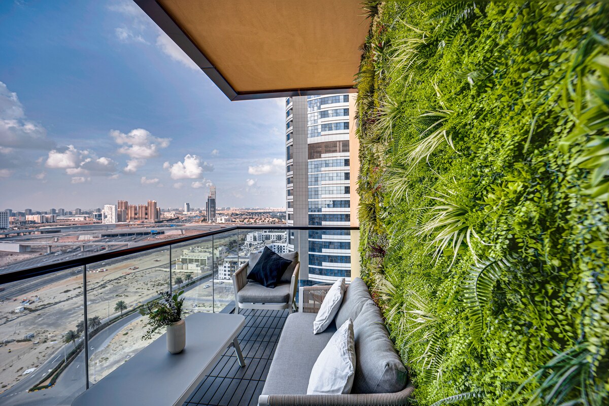 A green living wall adorns the balcony, providing a vibrant contrast against the modern building architecture. Comfortable seating is arranged on a wooden deck, offering an inviting space to relax while enjoying expansive views of the surrounding city and landscape.