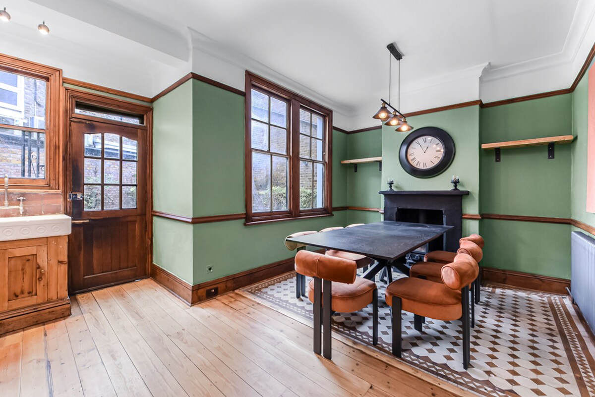 The dining area is illuminated by natural light filtering through large windows, highlighting the green walls and warm wooden elements. A dark table is surrounded by leather chairs, with a clock mounted above a decorative fireplace, enhancing the inviting ambiance.