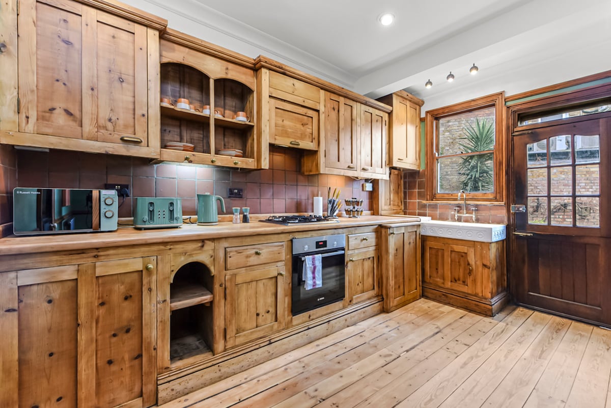 A spacious kitchen is presented with wooden cabinetry and a natural wood floor. Modern appliances, including a microwave and oven, are integrated within the design. A window offers natural light, revealing a view of greenery outside. A sturdy wooden door provides access to the courtyard.