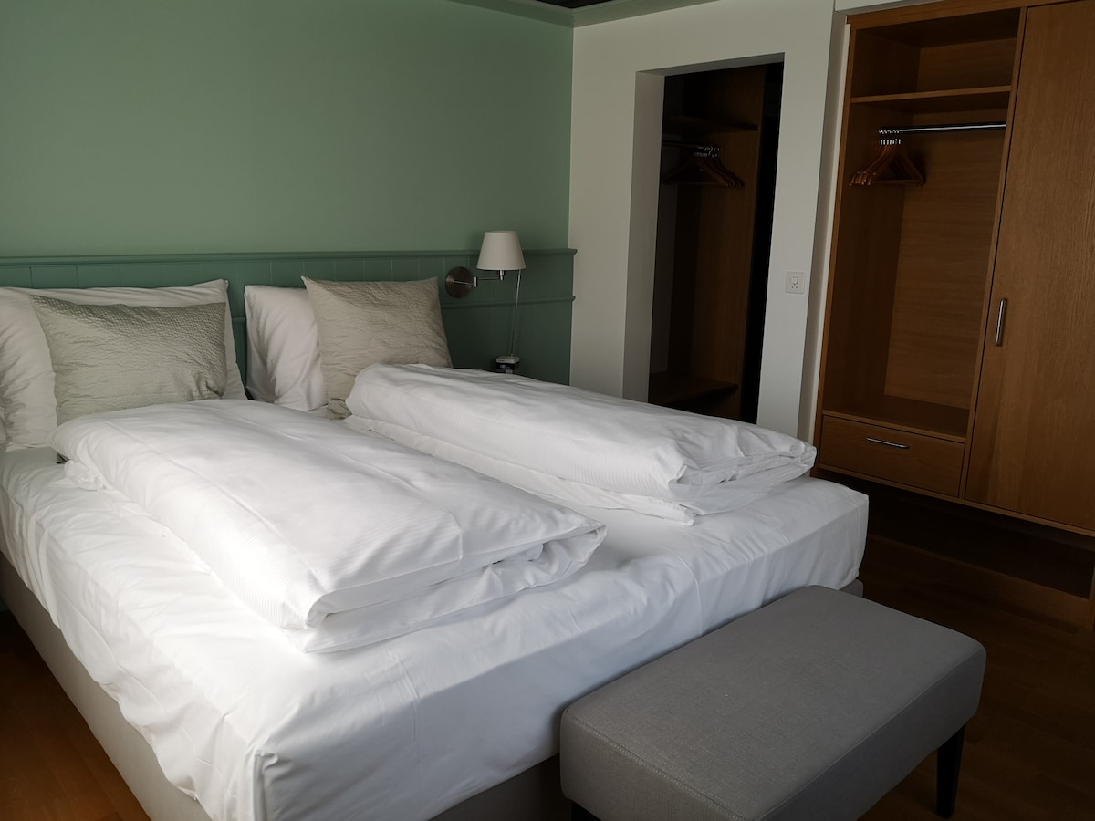 A well-lit bedroom features a spacious bed with neatly arranged white bedding and two sets of pillows. A minimalist bedside lamp stands beside the bed, which is paired with a gray upholstered bench. A wardrobe is visible, showcasing wooden hangers and a sleek design.