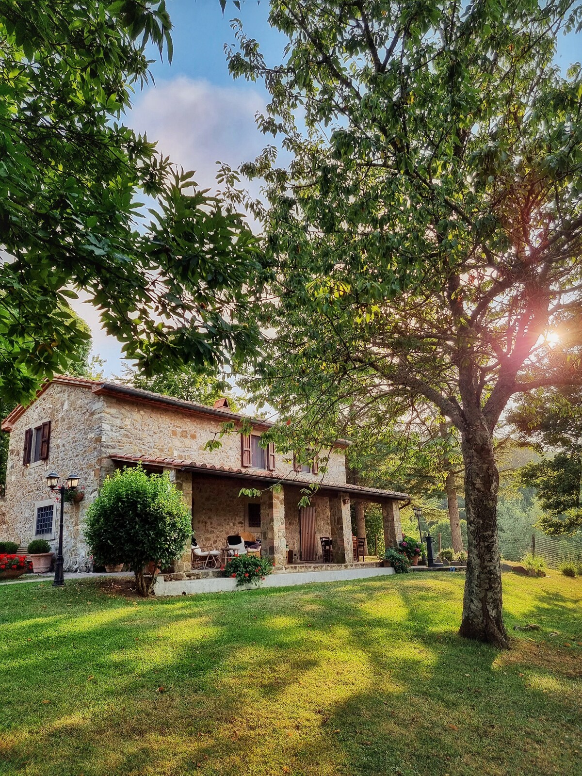 A stone villa is surrounded by lush greenery, featuring a spacious porch with seating arrangements. Sunlight filters through the leaves, creating dappled shadows on the manicured lawn. Large windows with shutters reflect the natural beauty of the peaceful setting.