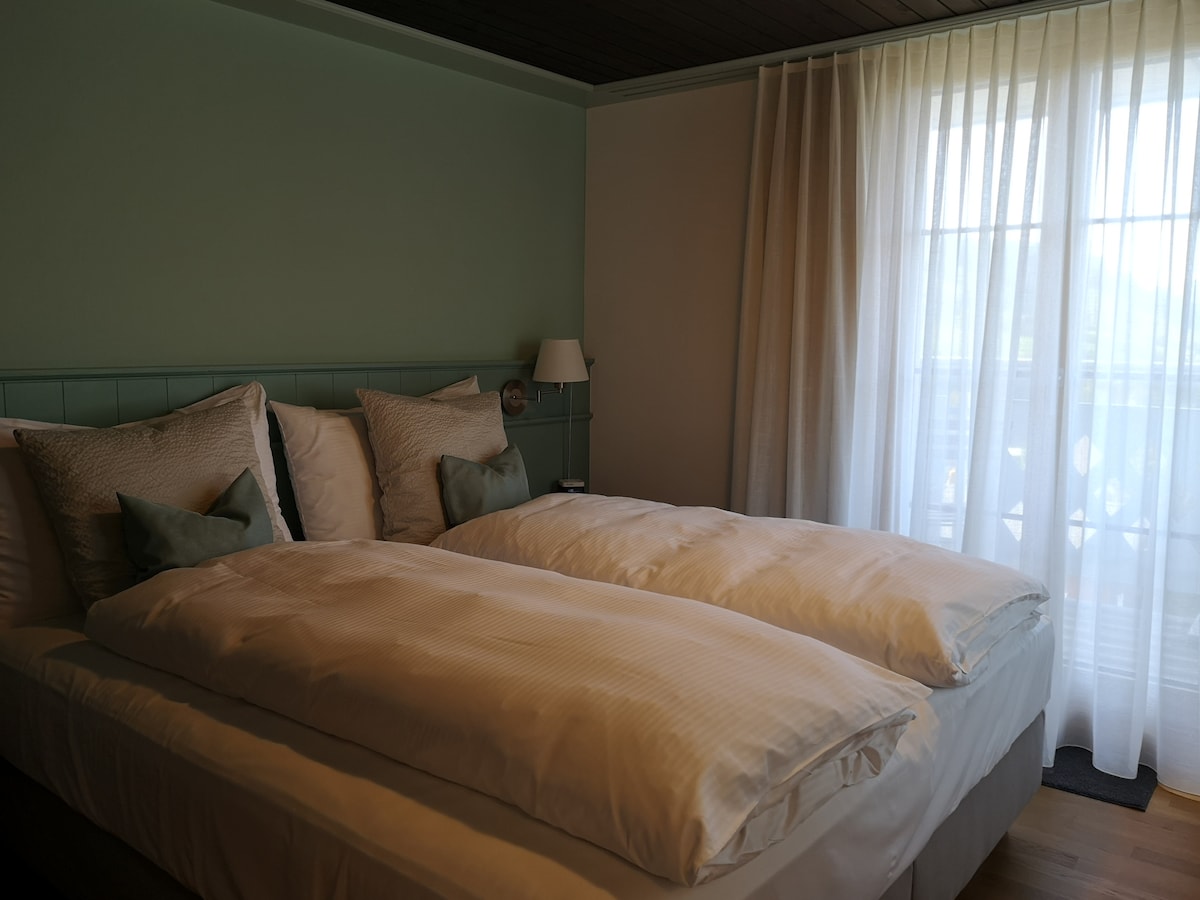 A serene bedroom features two spacious beds dressed with crisp white linens and soft, decorative pillows. Natural light filters through sheer curtains, illuminating the soothing green accent wall and enhancing the tranquil ambiance.