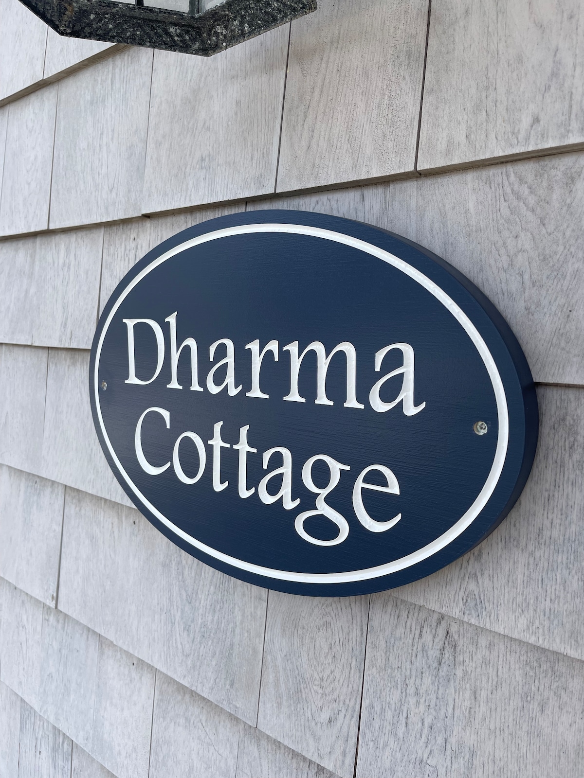 A circular wooden sign displaying the name 'Dharma Cottage' is mounted on the exterior wall of the cottage. The dark blue background contrasts with the white lettering, providing clear visibility. The textured wood siding of the cottage serves as a backdrop, enhancing the sign's presence.
