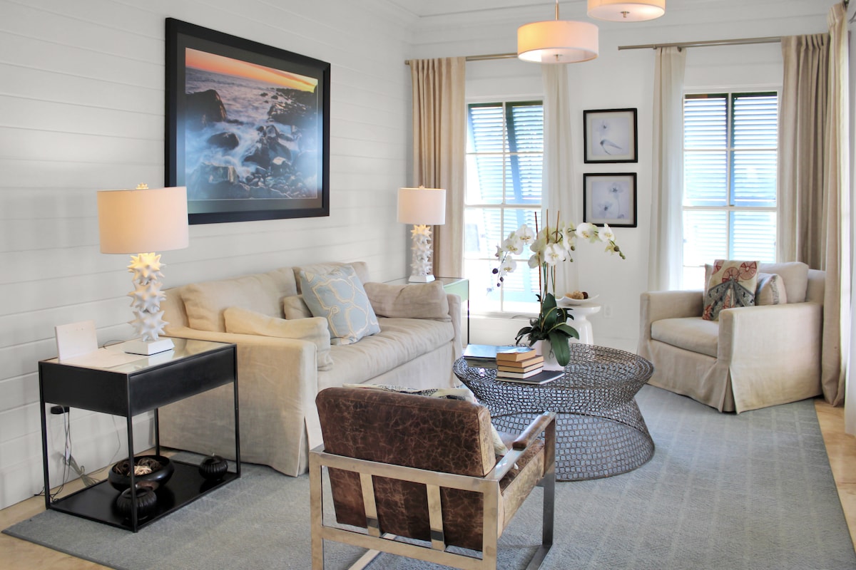 A bright living area features a light-colored sofa paired with a comfortable armchair. A round coffee table sits at the center, surrounded by soft area rugs. Large windows allow natural light to fill the space, pairing well with the neutral decor and artwork on the walls.