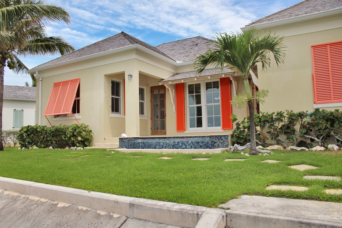 A single-story house features vibrant orange shutters and large windows. A small patio is visible, framed by lush greenery and decorative stones. A pathway leads to the entrance, adding to the inviting appeal of the exterior.