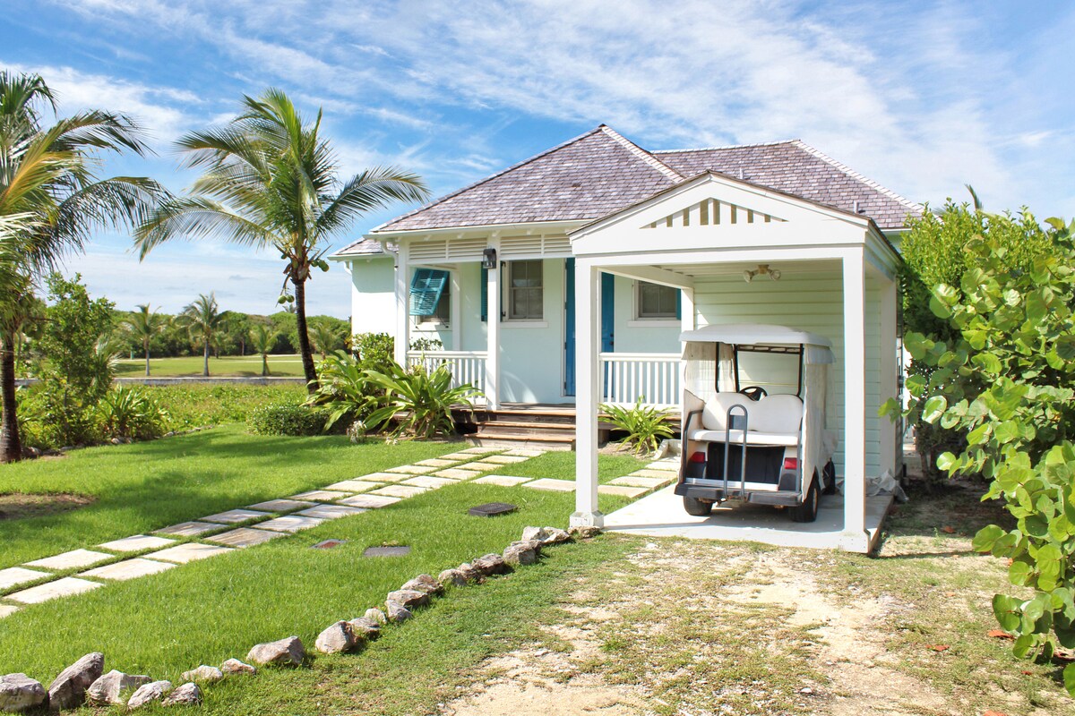 A charming cottage is surrounded by lush green landscaping, featuring a stone pathway leading to the front porch. A covered area houses a golf cart, with palm trees swaying gently in the background under a clear blue sky.