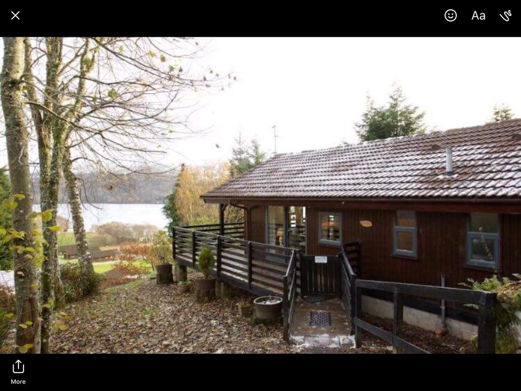 A contemporary lodge is nestled among trees, featuring a sloped pathway leading to a covered entrance. The wooden exterior complements the natural surroundings, while expansive windows offer views of the lake in the background. Seasonal foliage adorns the ground, adding a touch of nature.