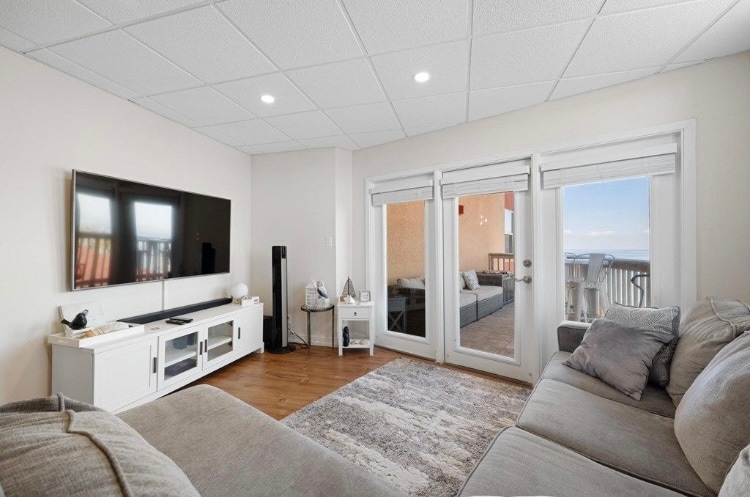 A cozy living area is presented with a spacious gray sofa and a flat-screen television mounted on the wall. Light is provided by recessed fixtures in the ceiling, and sliding glass doors lead to a balcony with views of the surroundings.