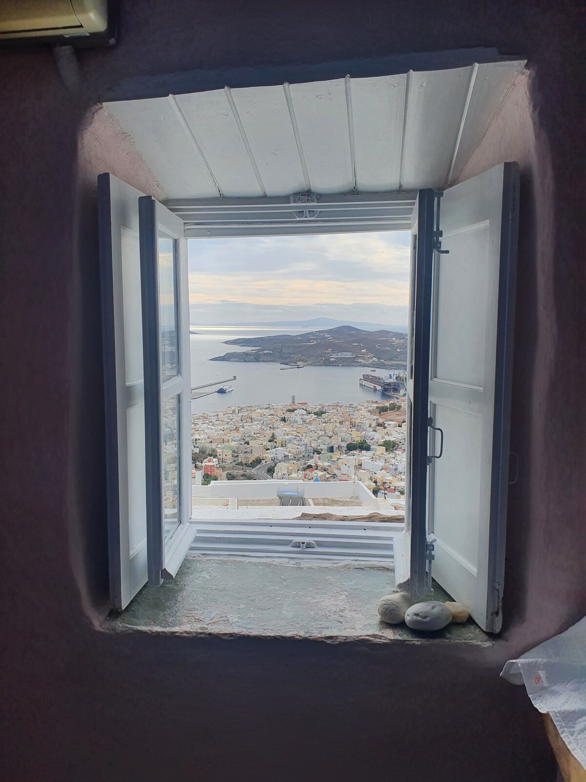 A window with open shutters reveals a stunning view of the coastline and the town below. The sea glistens in the distance, while the rooftops of colorful buildings create a picturesque landscape. The frame of the window is made from traditional stone, adding to the charm.