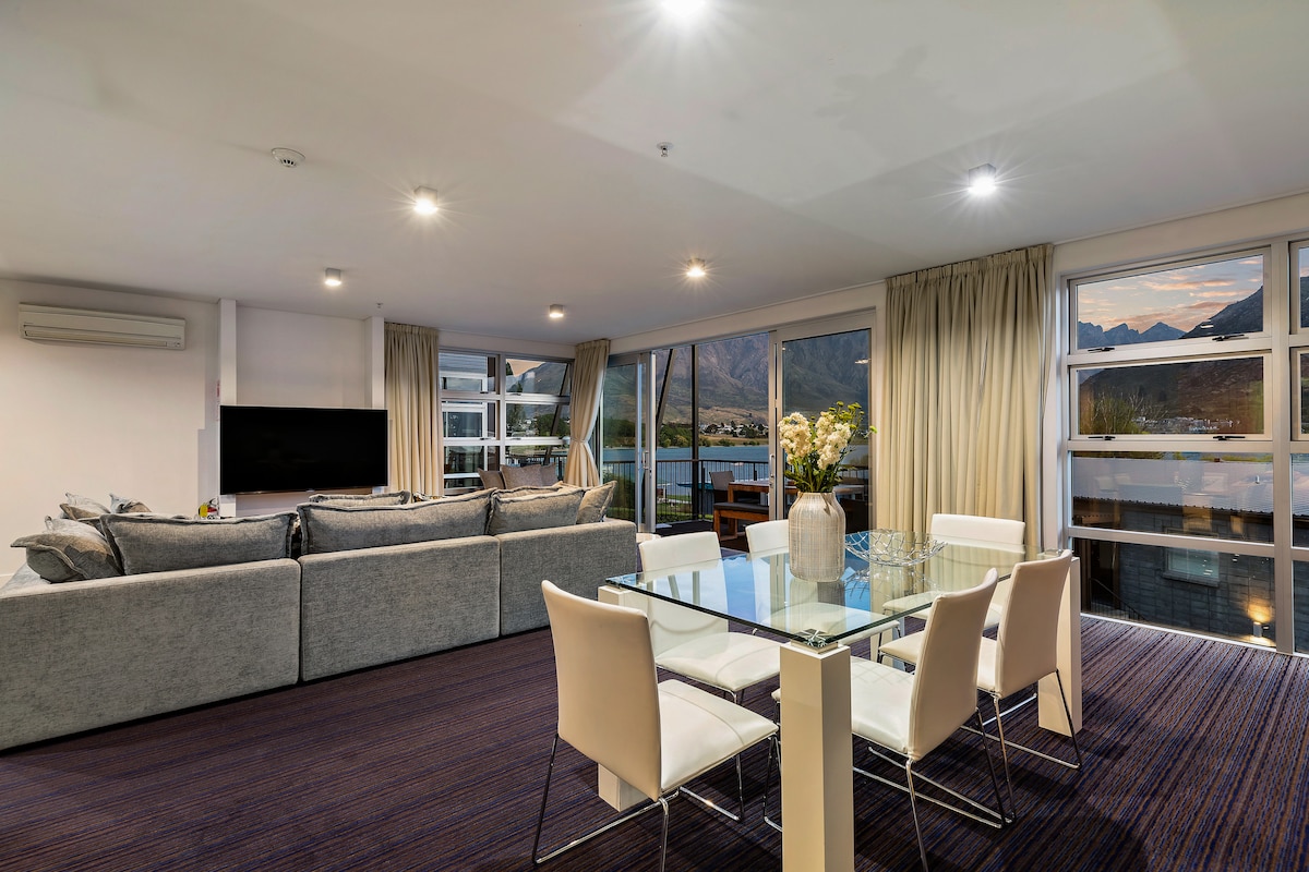 A light-filled lounge area features comfortable seating arranged around a large television, with expansive windows providing views of the landscape. A dining table with white chairs is positioned nearby, complementing the modern decor and inviting ambiance of the space.