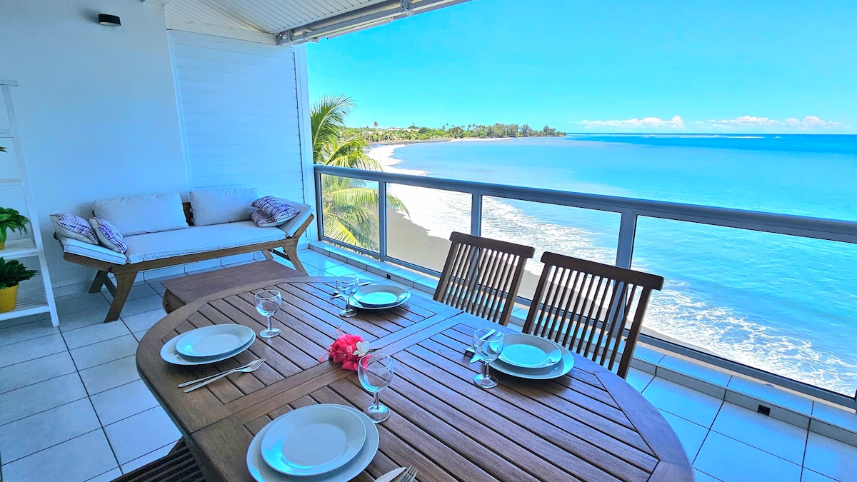A spacious terrace features a wooden dining table set for six, with plates and glasses neatly arranged. A comfortable seating area is visible, framed by palm trees and offering a clear view of the tranquil ocean and shoreline.