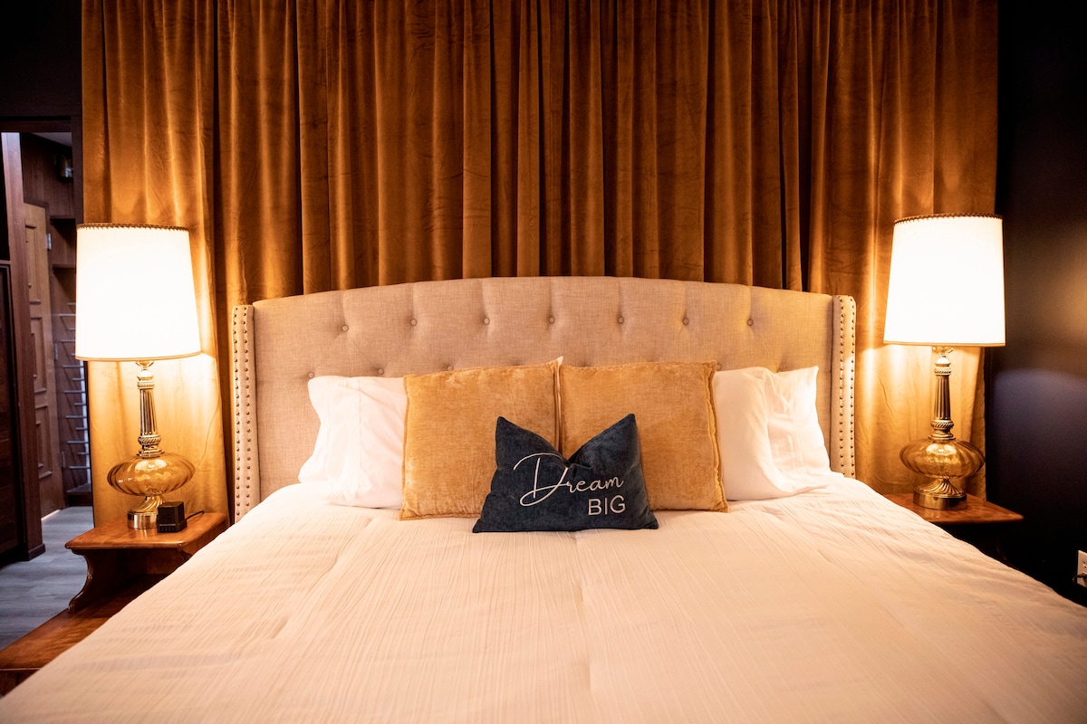 A spacious bed is framed by a soft upholstered headboard, complemented by elegant lamps on either side. Golden curtains add a touch of warmth, while a decorative pillow with the text 'Dream BIG' enhances the inviting ambiance of the room.