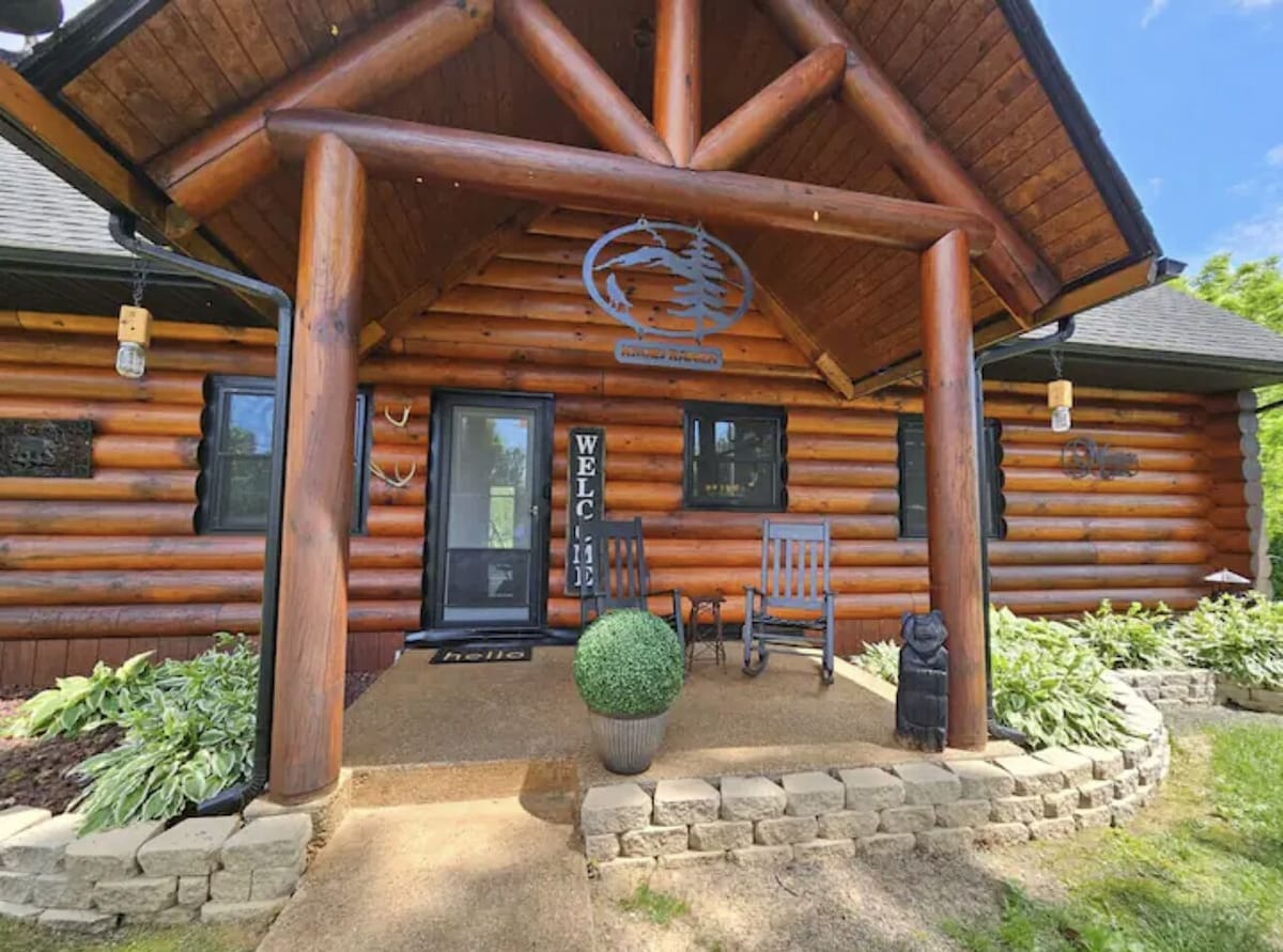 The entrance showcases a log cabin facade with rustic wooden beams. A welcoming mat is positioned in front of a black door, flanked by two rocking chairs. Lush greenery and decorative elements enhance the inviting outdoor space.