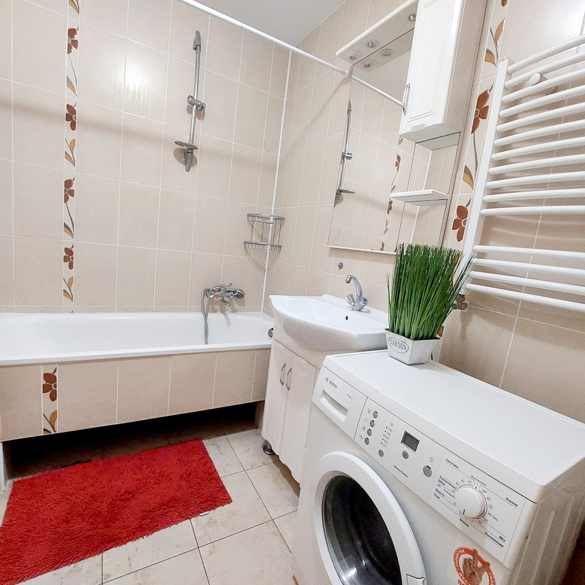 A neutral-toned bathroom features a bathtub with a shower attachment, accompanied by a sink and a washing machine. Wall shelves hold a small plant. A vibrant red rug lies in front of the bathtub, adding a pop of color to the space.