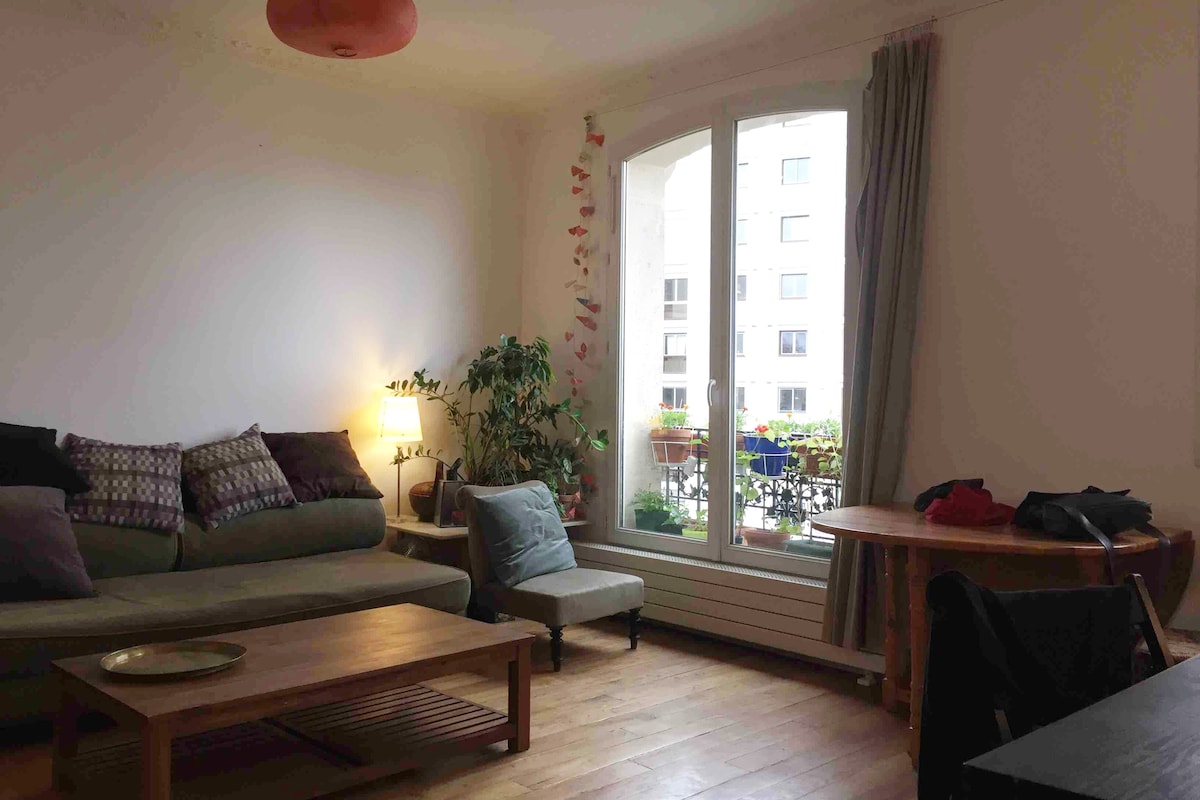 A bright living area features large windows overlooking a balcony with colorful plants. A cozy sofa with assorted pillows sits next to a small green armchair. A wooden coffee table and dining table complement the warm wooden flooring. Soft lighting is provided by a table lamp.