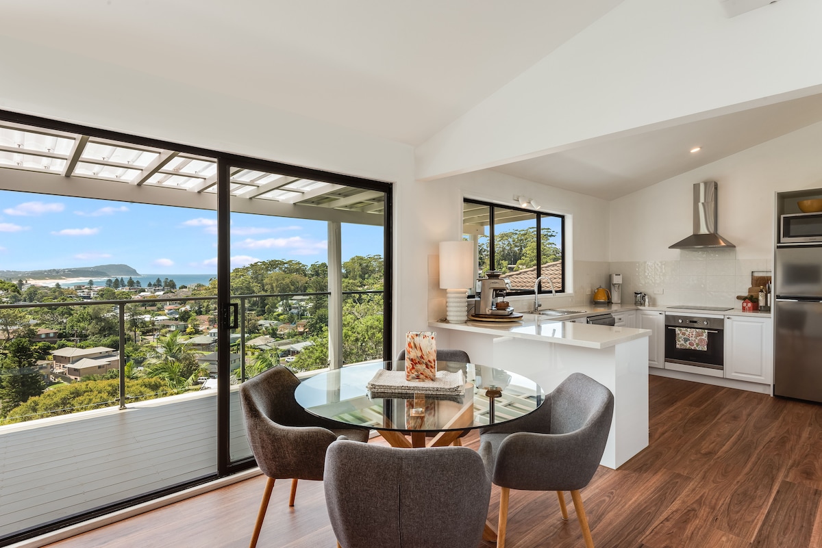 A modern kitchen and dining area feature large windows offering views of the ocean and lush greenery. The round dining table is set with simple decor, while sleek appliances complement the spacious layout. A well-lit atmosphere is created by natural light flowing through the open space.