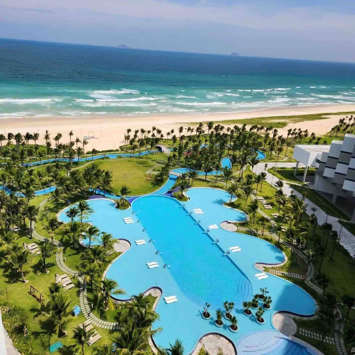 An expansive view of the resort's swimming pool is showcased, featuring a unique curved shape surrounded by lush greenery. The pool is bordered by lounge chairs, with the beach and ocean visible in the background, complemented by clear blue skies.