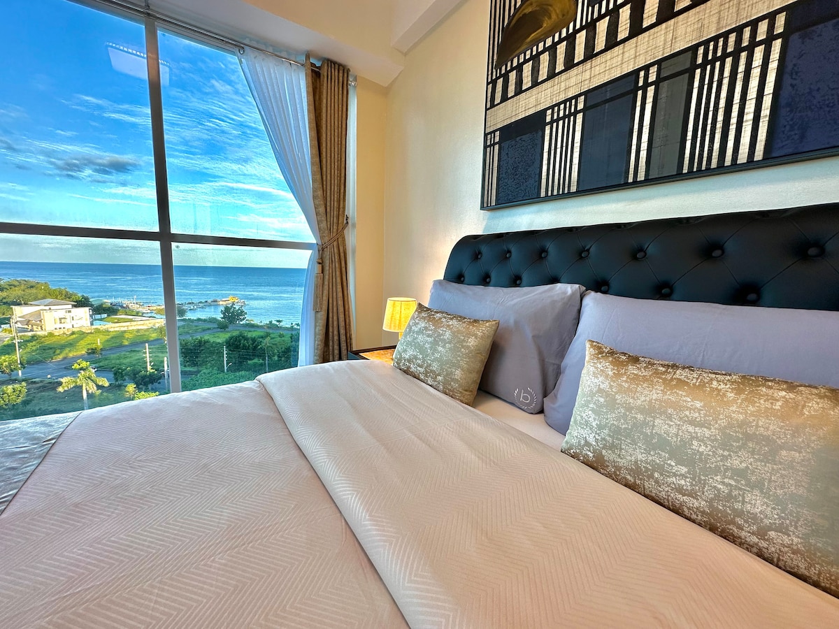 A spacious bed is positioned in front of a large window offering views of the ocean and surrounding landscape. Soft linens and decorative pillows complement the design, while natural light illuminates the room, creating a peaceful ambiance.