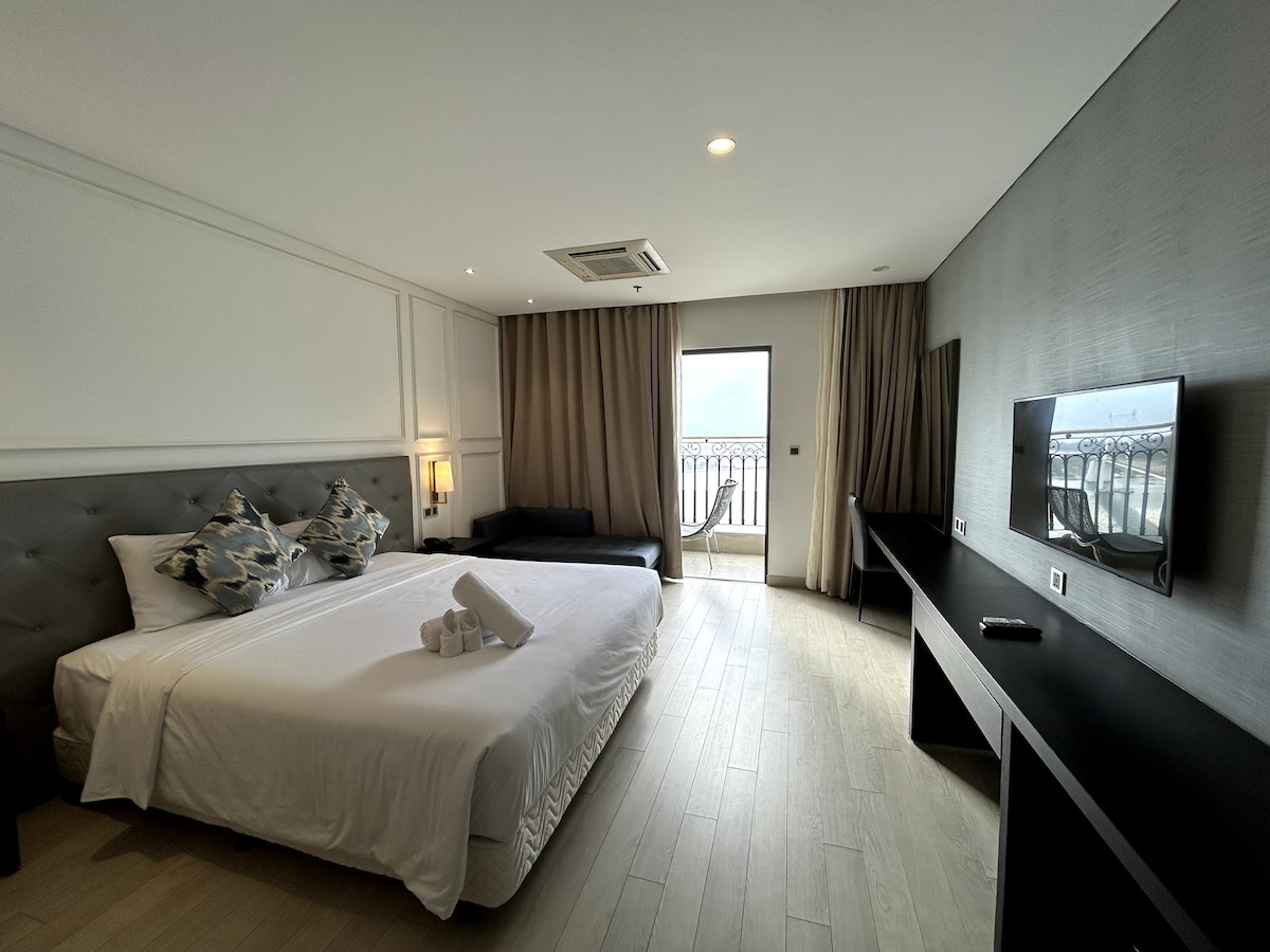 A spacious room features a large king bed with decorative pillows. A neutral color palette is complemented by soft natural light from large windows leading to a private balcony. A sleek desk and a sofa can be seen, enhancing the room's functionality.