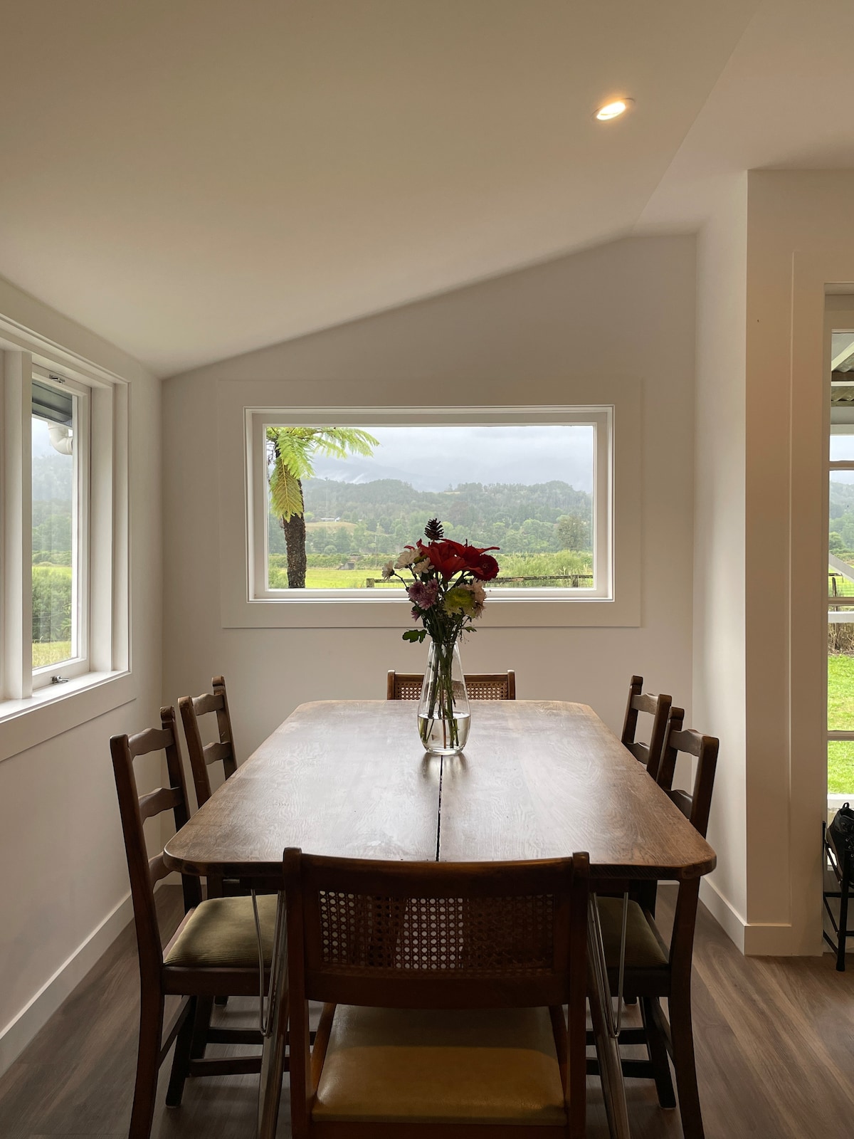 A spacious dining area features a large wooden table surrounded by eight chairs. A vase with fresh flowers sits at the center, while a window offers views of rolling greenery and distant mountains. Natural light brightens the room, creating an inviting atmosphere.