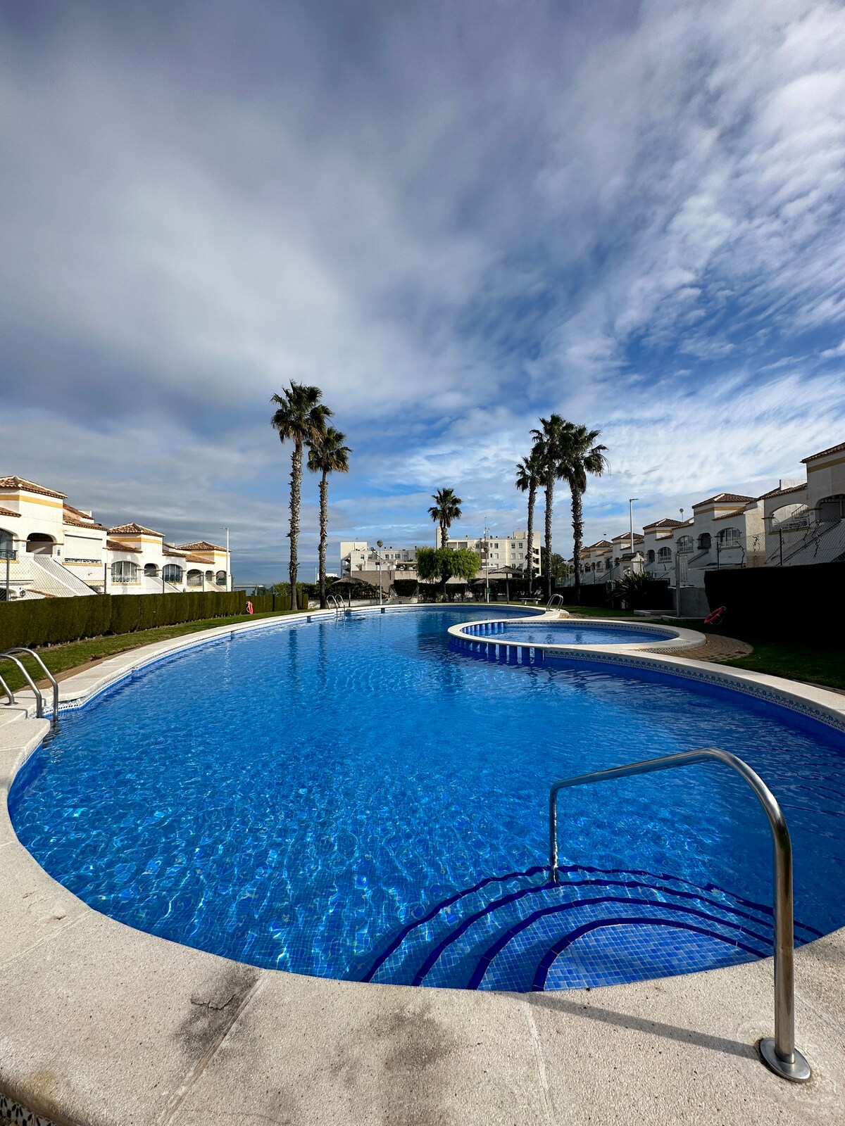 A large swimming pool is framed by palm trees, with clear blue water reflecting the sky. Steps lead into the pool from the surrounding area, while a smaller circular pool is visible alongside. The backdrop features residential buildings, enhancing the tranquil setting.