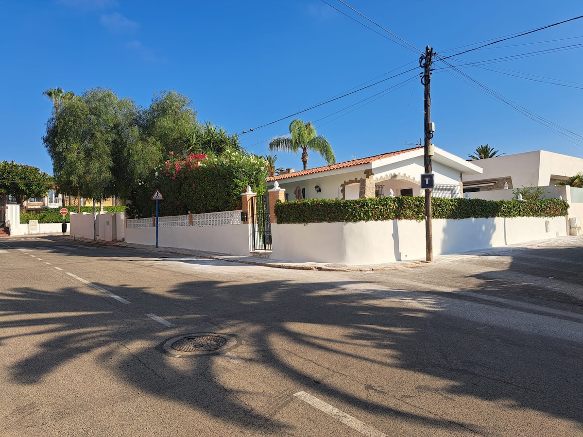 A corner view of a sunny residential property is presented, featuring a white-walled exterior with lush green shrubs and palm trees. The tranquil street is lined with additional greenery, and the clear blue sky adds to the serene atmosphere surrounding the home.