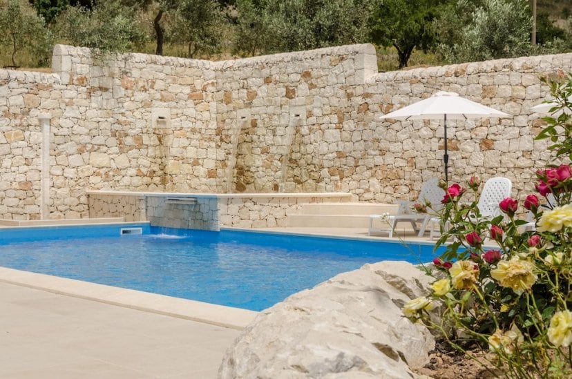 A serene swimming pool is encircled by rustic stone walls, creating a tranquil atmosphere. Sun loungers are positioned near an umbrella, while vibrant roses add a pop of color to the environment. Water cascades gently from the wall, enhancing the peaceful setting.