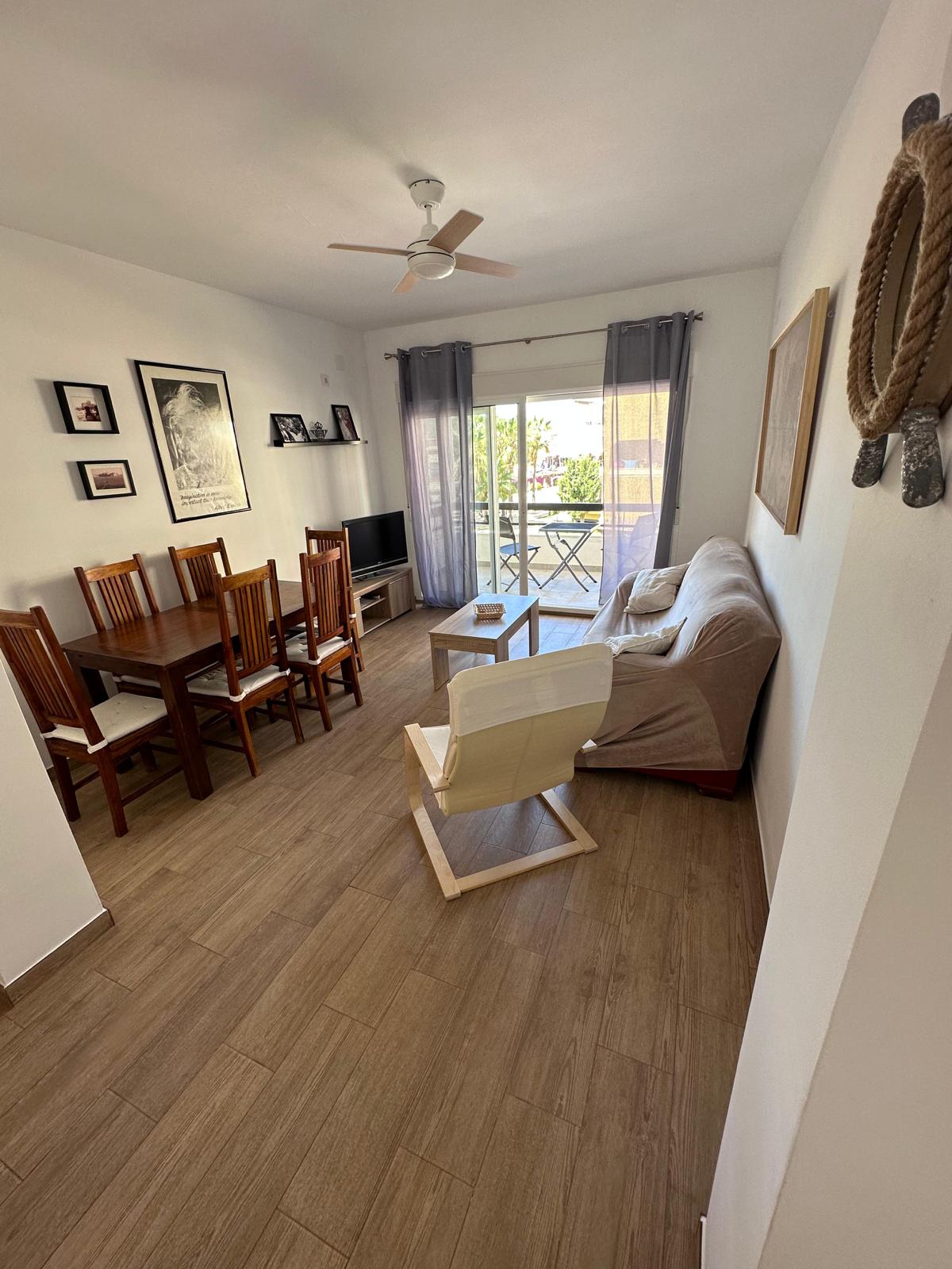 A living area features a dining table with eight wooden chairs, along with a comfortable sofa and a light-colored armchair. Large glass doors open to a terrace, allowing natural light to fill the space. Decorative wall art adds character to the room.