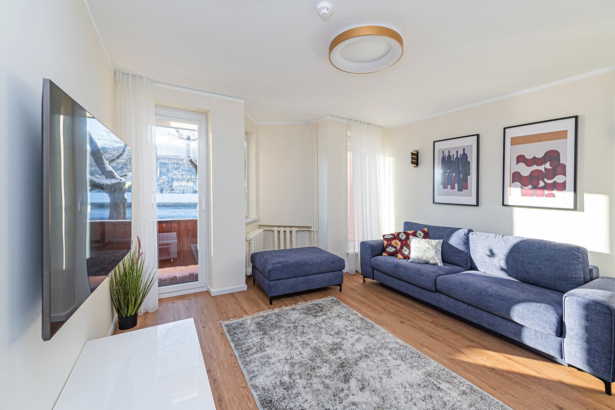 The living room features a comfortable blue sofa with a chaise lounge, positioned next to a large wall-mounted TV. Natural light enters through the window, which has sheer curtains, illuminating the stylish decor. A soft area rug adds warmth to the wooden floor.