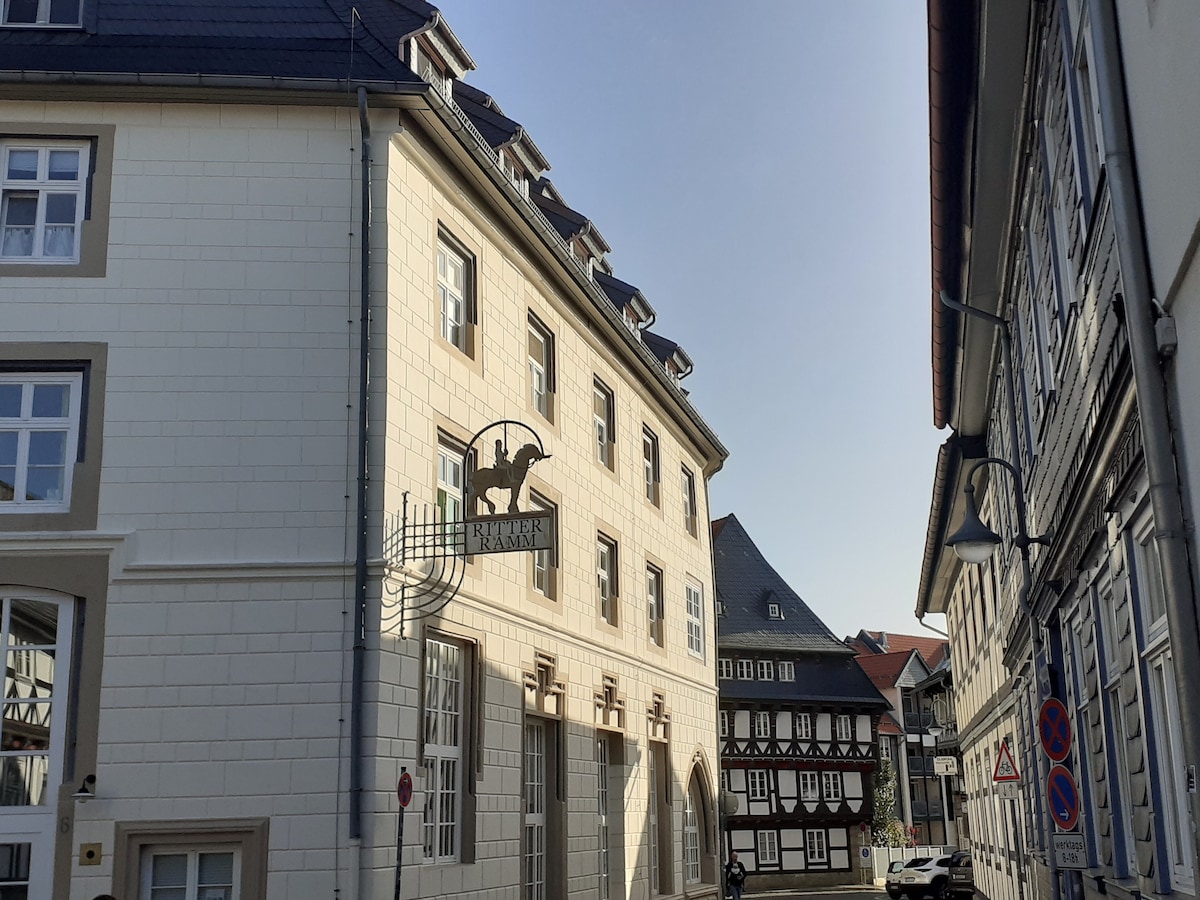 The exterior of a historic building is shown, featuring a distinct sign hanging from the facade. Surrounding structures exhibit traditional architecture, with houses displaying timber framing. Natural light illuminates the scene, highlighting the clean lines and contrasting materials of the buildings.