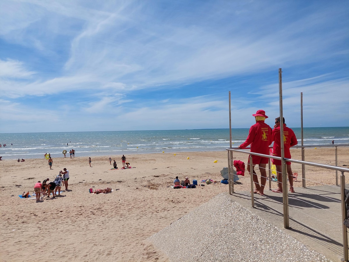 Two lifeguards dressed in red uniforms stand watch over a sandy beach, with gentle ocean waves in the background. Groups of beachgoers enjoy the sun on towels, while children play in the sand. The sky is clear with wispy clouds.