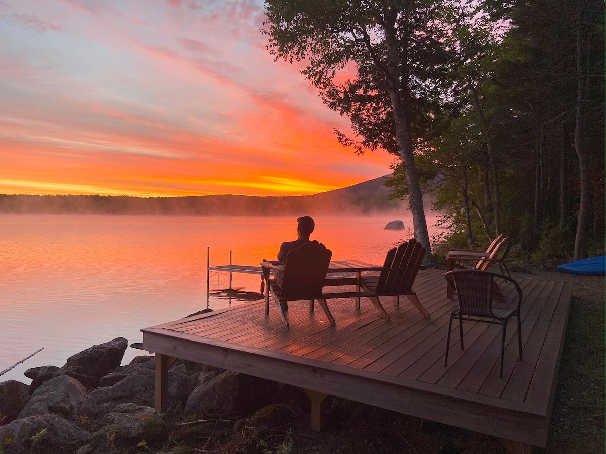 A tranquil lakeside deck is presented, featuring a variety of seating options including two Adirondack chairs. The vibrant sunset casts warm hues over the water, while mist gently rises in the background, framed by trees and a distant mountain.