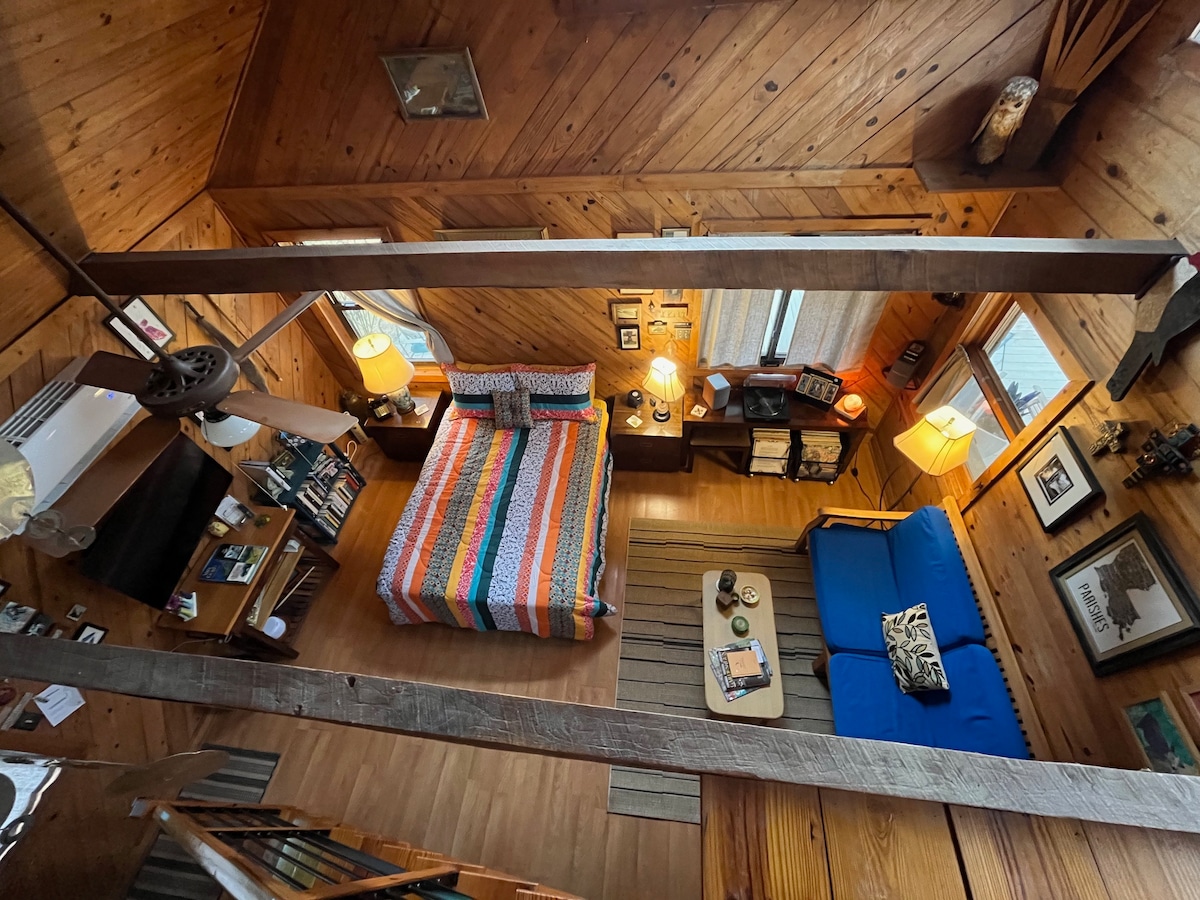 An overhead view of the open space design showcases a queen-sized bed with a colorful striped blanket. A twin-sized bed/sofa is positioned in the loft area. Wooden walls provide warmth, while two lamps illuminate the cozy interior. A seating area with a small table adds to the charm.