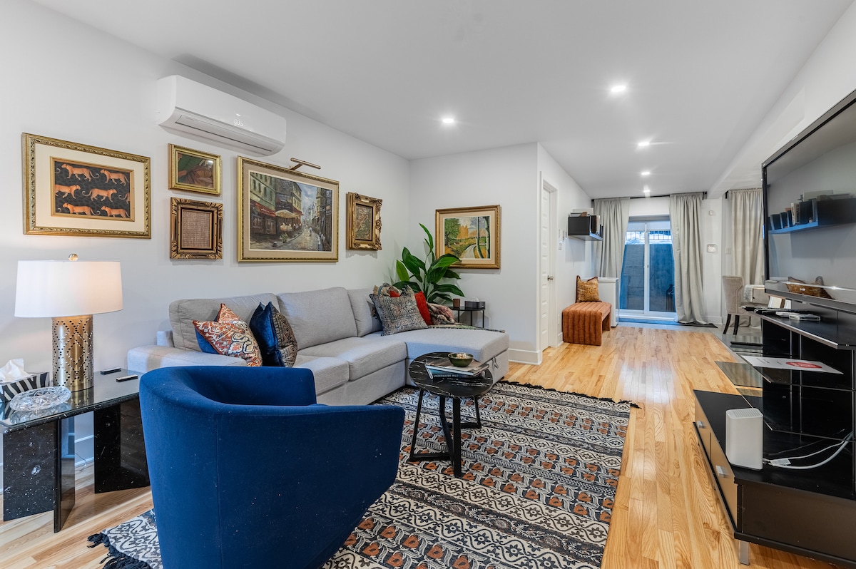 A serene living room features a grey sectional sofa complemented by a blue accent chair. Artwork adorns the walls, and a patterned rug covers the hardwood floor. A sleek black entertainment unit holds a large TV, while natural light enters through glass doors leading to an outdoor area.