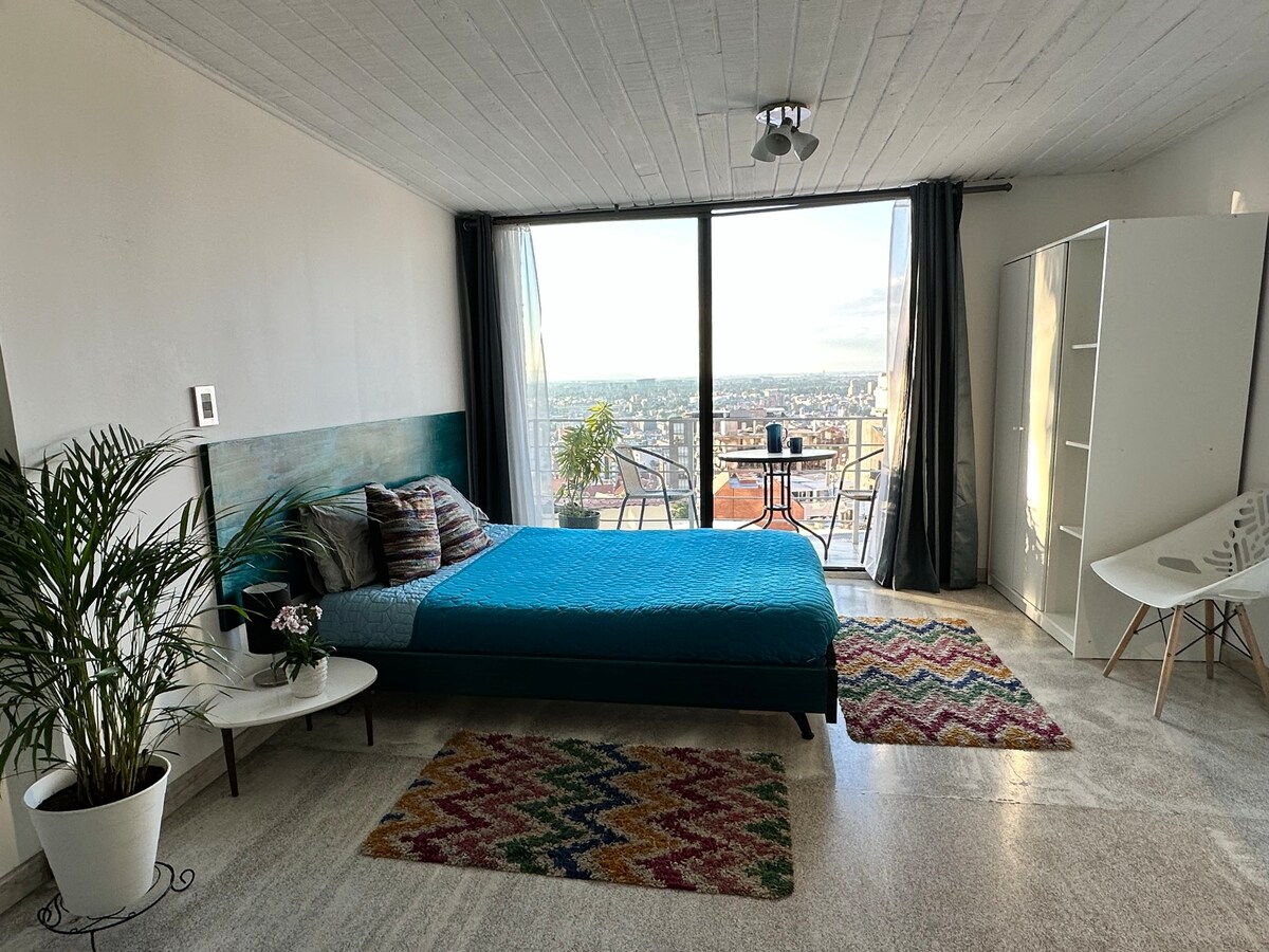 A spacious bedroom features a large bed with a vibrant blue coverlet. A small balcony with two chairs offers sweeping views of the city. Decorative rugs add color to the floor, while a potted plant provides a touch of greenery.
