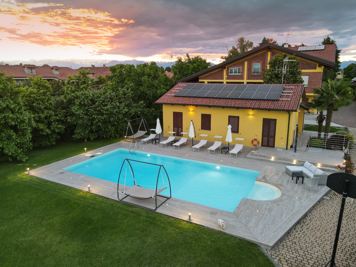 A spacious outdoor area features a rectangular pool surrounded by lounge chairs and umbrellas. A swing chair is positioned beside the pool. The building is painted yellow, has a red-tiled roof, and solar panels on top, with greenery enhancing the landscape.
