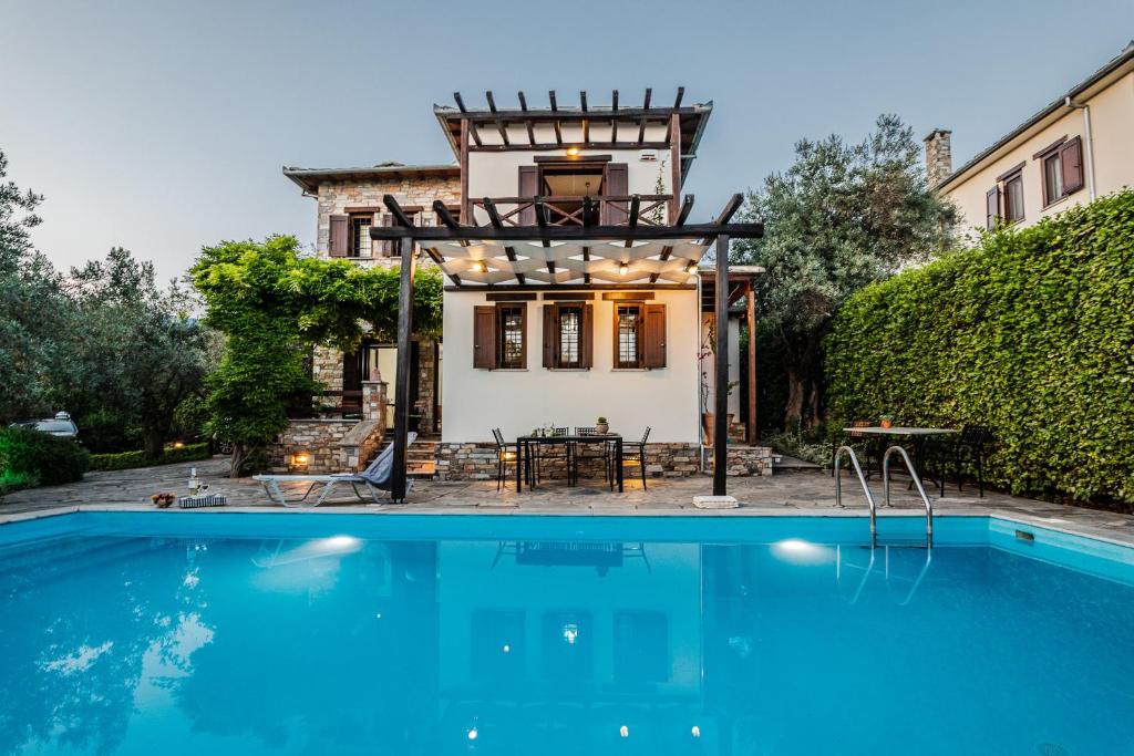 A serene pool area is framed by a stone pathway and lush greenery. The two-story house features wooden shutters and a patio with seating under a trellis adorned with lights. The inviting pool reflects the evening sky, creating a calming ambiance.