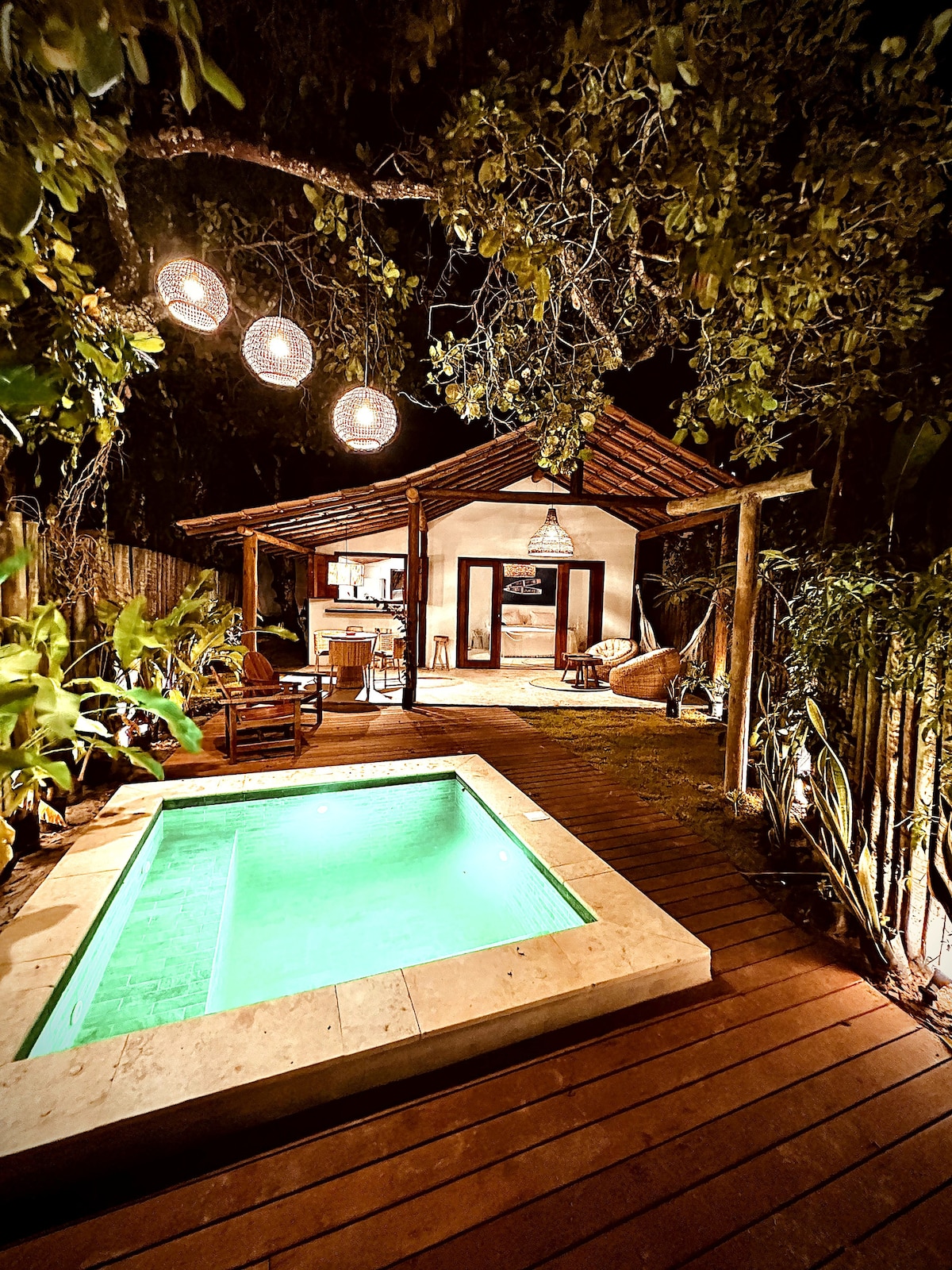A quaint outdoor area features a private green-tinted plunge pool, complemented by natural wood decking. Surrounding foliage creates a serene setting, while pendant lights hang from tree branches, illuminating the space. The cozy structure in the background houses an inviting seating arrangement.