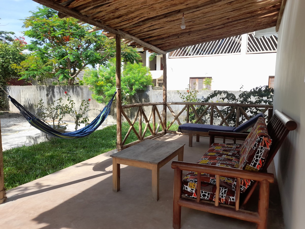 An outdoor terrace area features a wooden table and seating with vibrant patterned cushions. A hammock is seen hanging between the posts. Lush green plants and trees surround the space, creating a natural and tranquil setting.