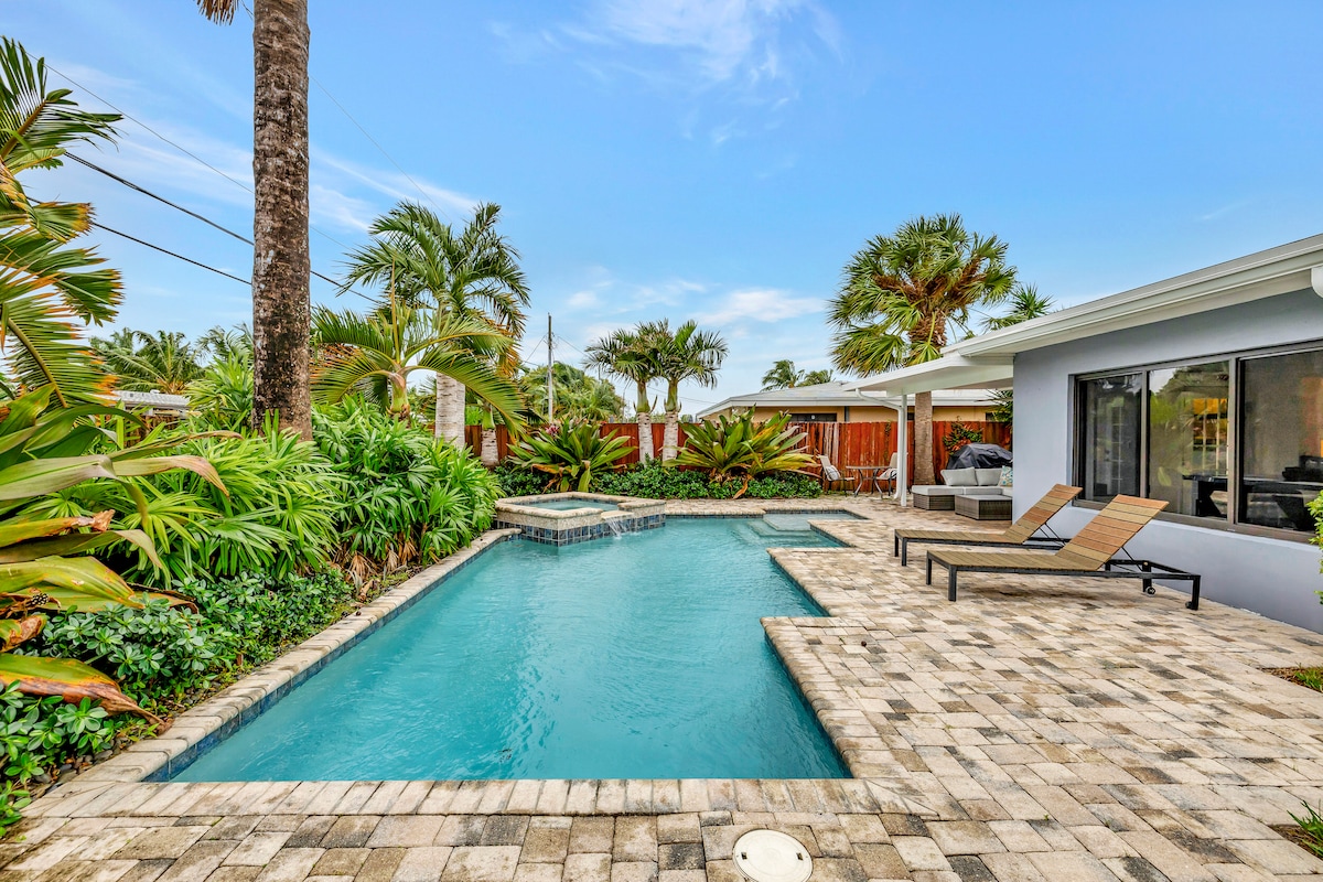 A landscaped outdoor area showcases a private saltwater pool surrounded by lush tropical greenery. Two lounge chairs are positioned beside the pool, and steps lead to a hot tub. The covered patio area offers shaded seating, enhancing the relaxing ambience of the backyard space.