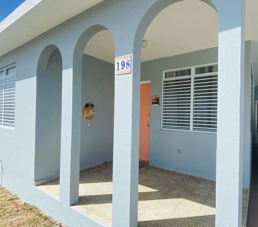 A welcoming entrance features a smooth stone floor and arched openings that lead to the inside. A house number is displayed on a blue wall beside a decorative sign. Light streams through large windows, enhancing the inviting atmosphere.