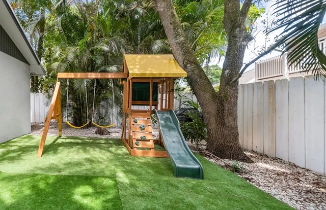 A wooden play structure is situated in a grassy backyard, featuring a slide and swings. Surrounding tall trees provide shade, while a gravelly area borders the space. The backdrop includes a white fence enhancing privacy.
