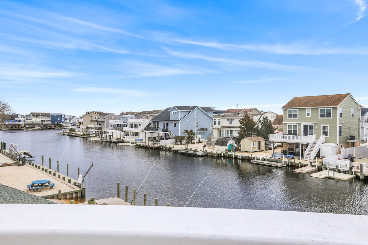A scenic view of a calm lagoon, with various colorful houses lining the water's edge. Docked boats are visible, alongside well-maintained docks, creating a relaxed coastal atmosphere. The sky is clear, enhancing the pleasant waterfront setting.