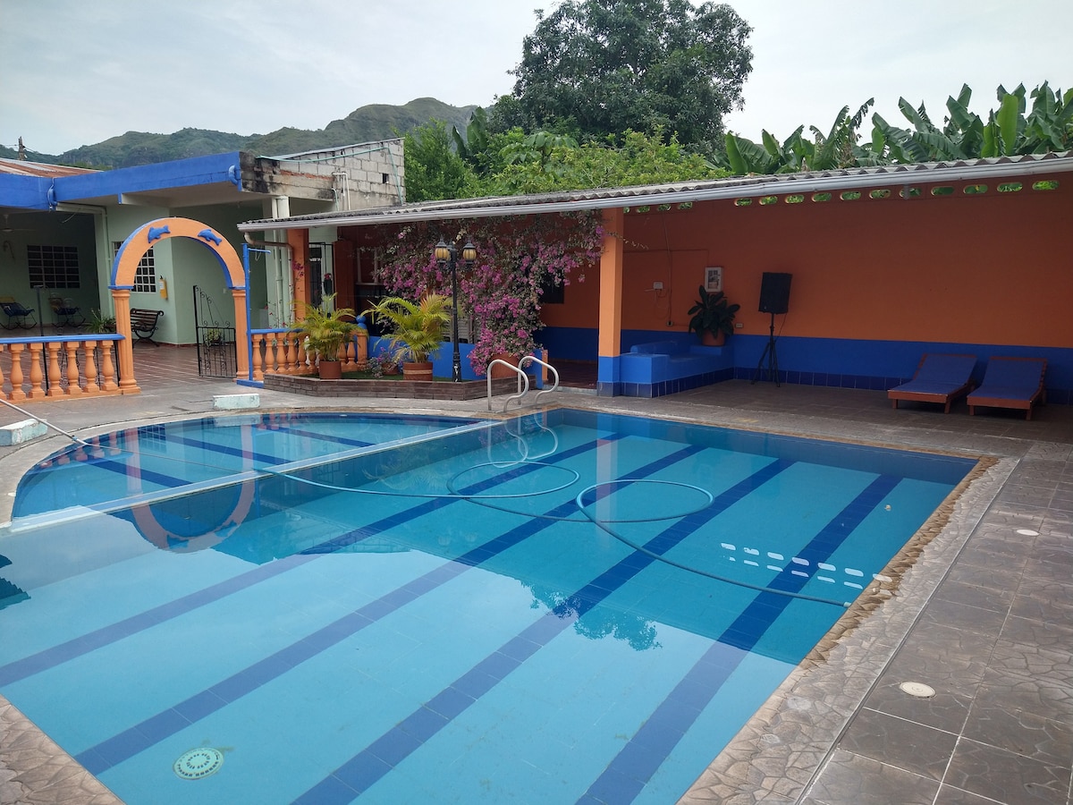 A clear swimming pool reflects surrounding colors, featuring a shallow area marked by light blue tiles. Lounge chairs line the opposite side, with vibrant walls decorated in orange and blue. Lush greenery and pink flowering plants border the space, enhancing the serene ambiance.