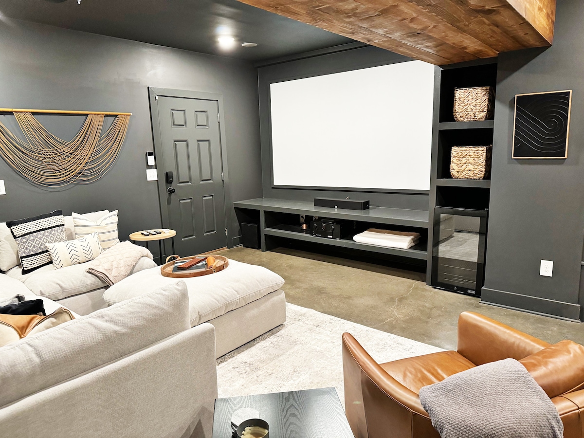 A modern basement theater area features a large projector screen mounted on a dark wall. Comfortable seating includes a plush sectional and leather chairs. Decorative accents include woven wall art and organized shelves, creating a cozy environment for movie nights or gatherings.